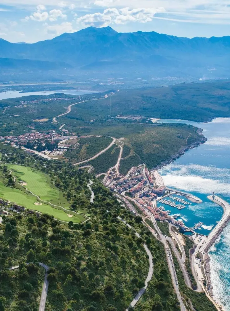 Pogled iz vazduha na obalu, naselje i marinu sa planinskim pejzažem u pozadini.