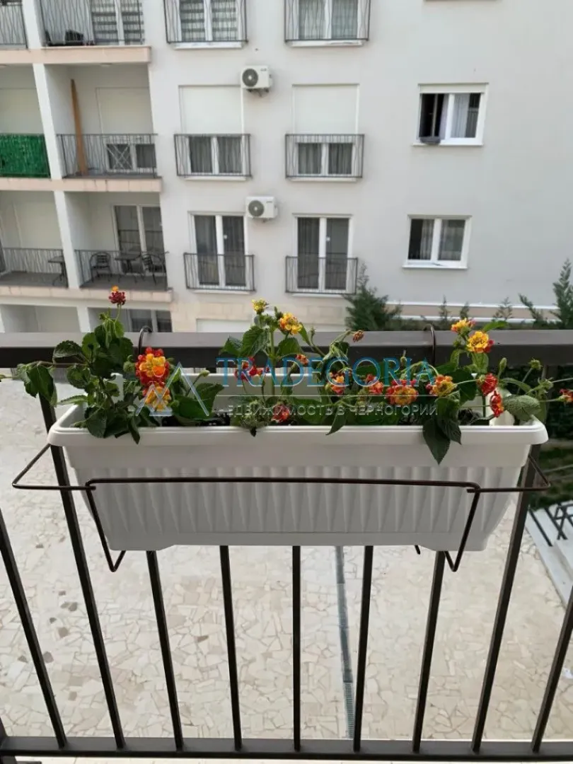 Pogled sa balkona sa žardinjerom i cvijećem prema zgradi.