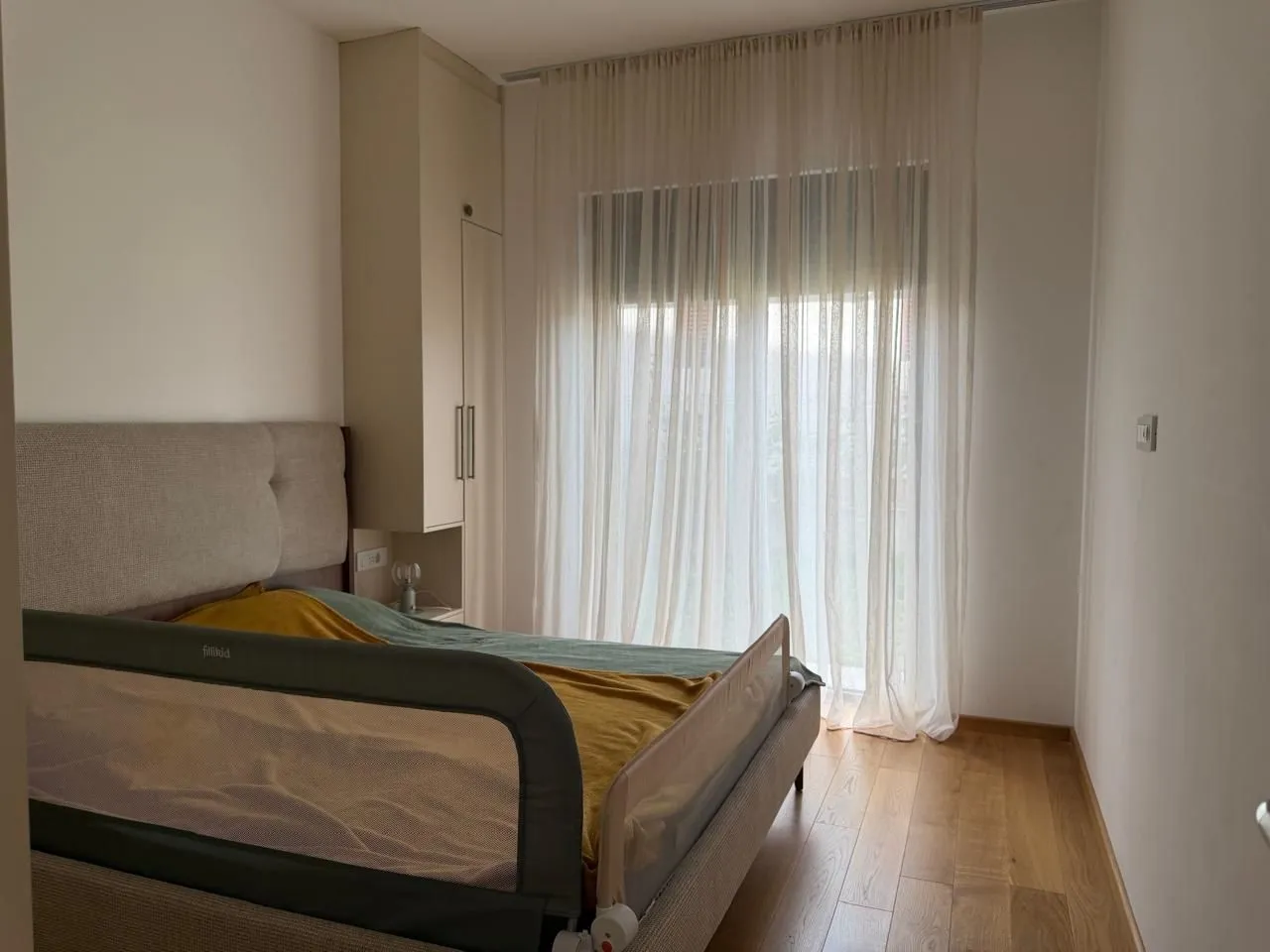 Namještena spavaća soba sa bračnim krevetom, ugradnim ormarom i balkonskim vratima sa bijelim zavjesama.