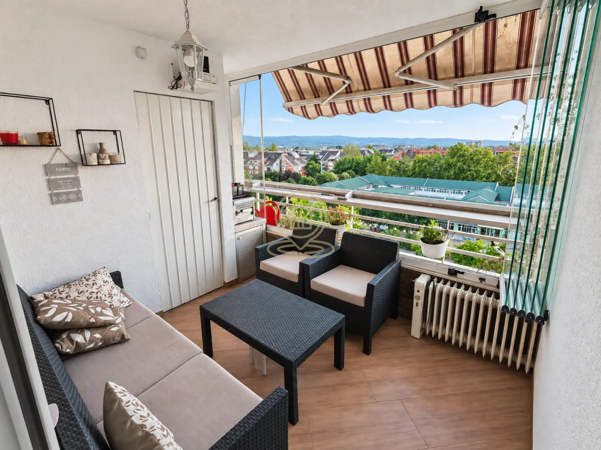 Zastakljeni balkon sa garniturom za sedenje i pogledom na naselje.