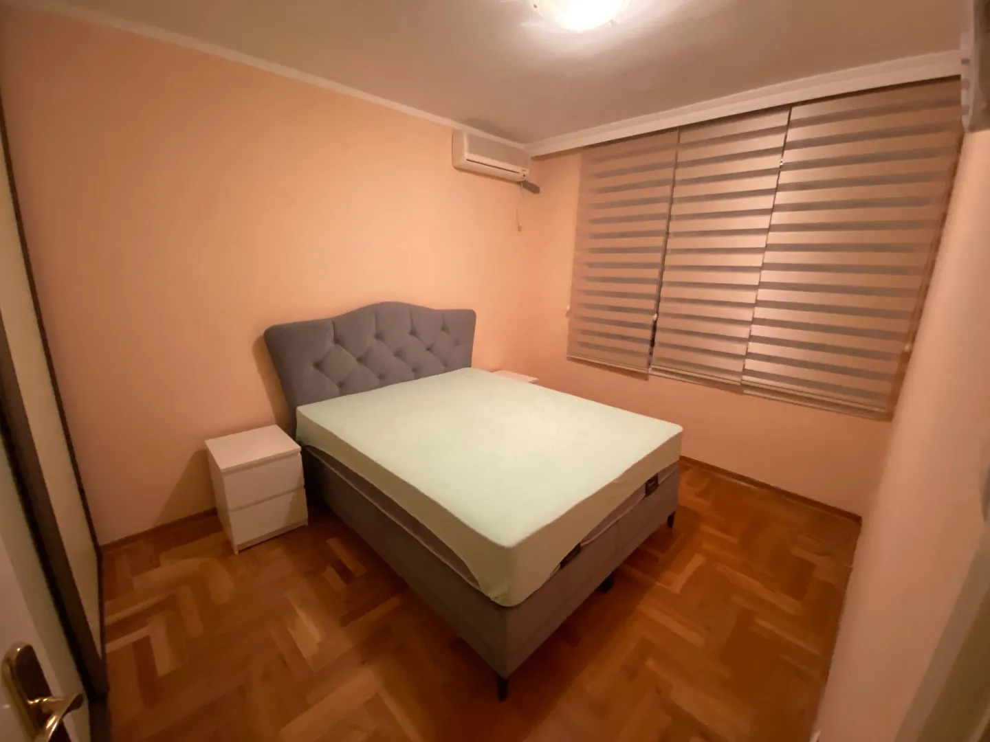 Namještena spavaća soba sa bračnim krevetom, noćnim stolićem i prozorom sa roletnama.