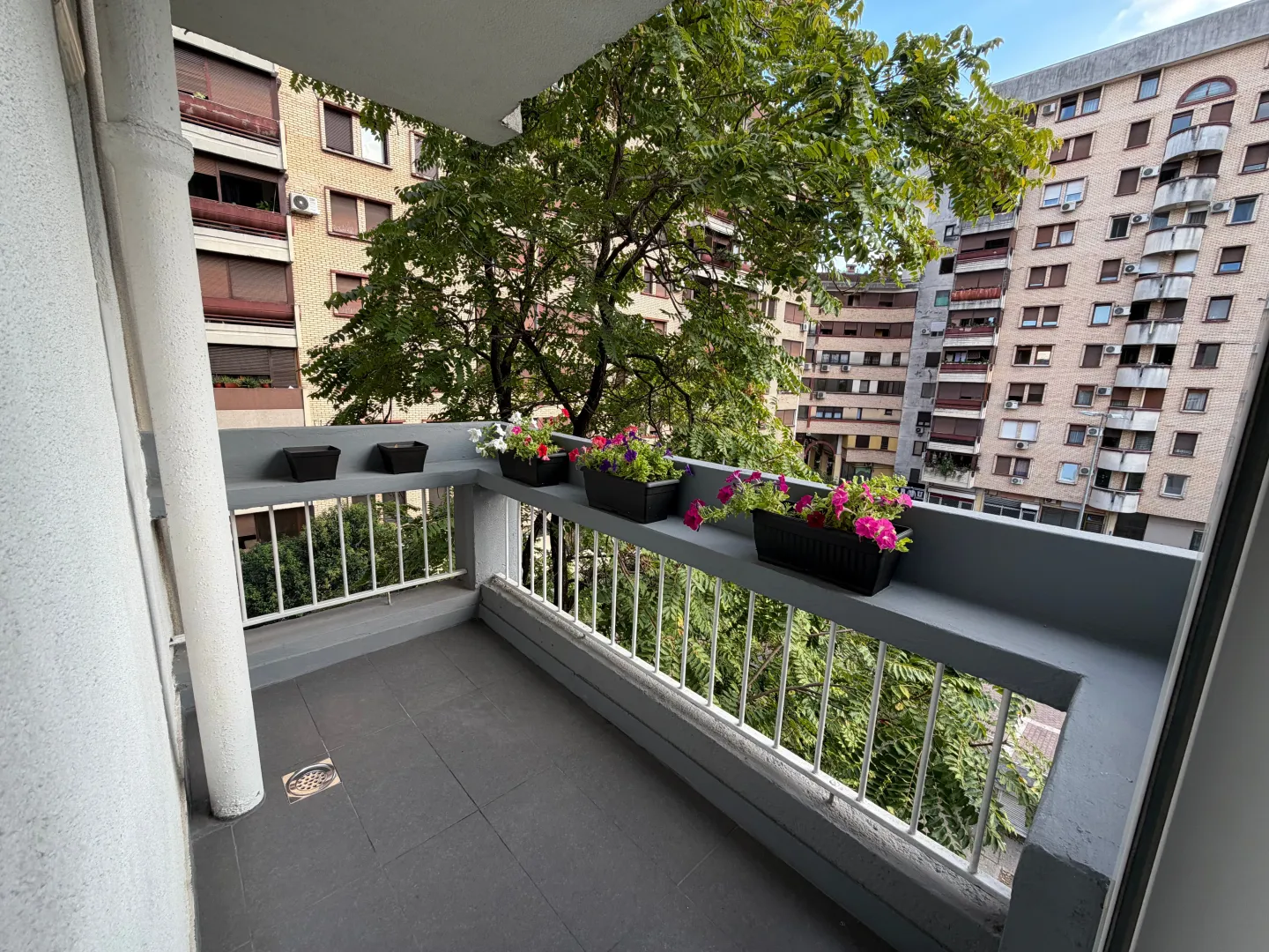 Manji uređeni balkon sa ogradom i saksijama cvijeća, sa pogledom na zgrade i drveće.