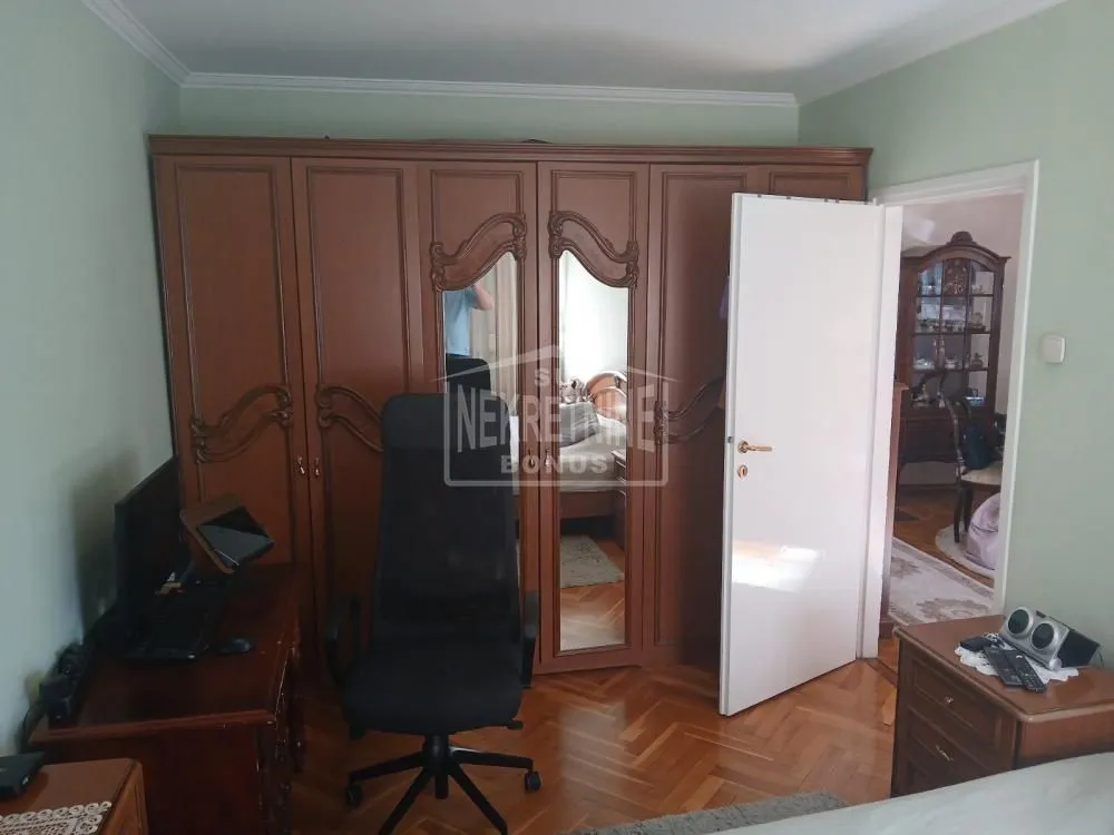 Spavaća soba sa velikim drvenim ormanom, radnim stolom i parketom na podu.