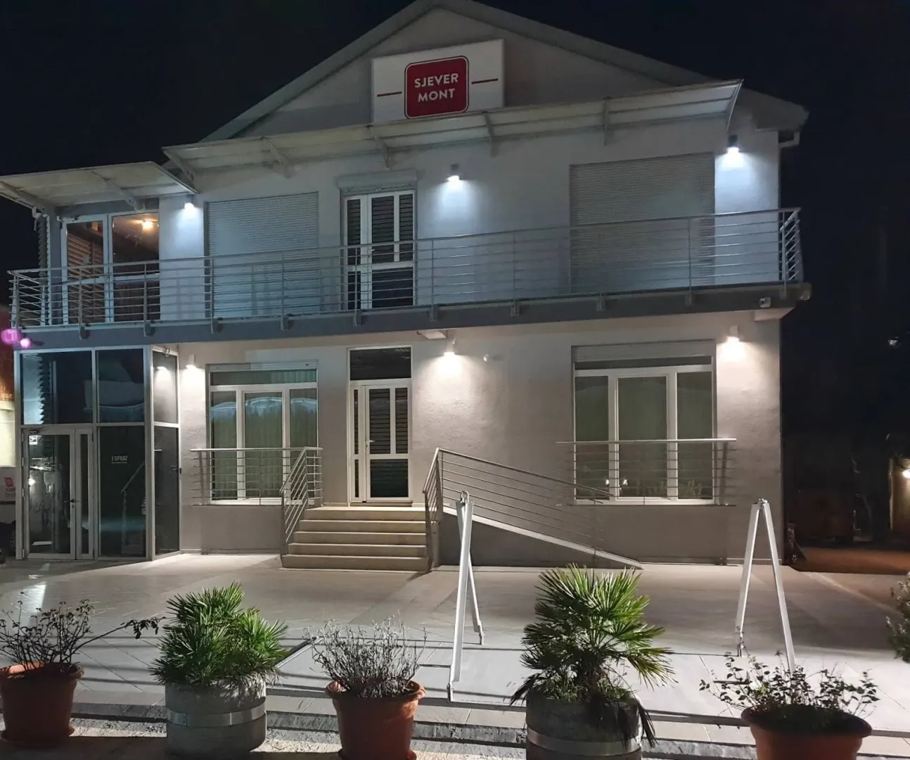 Exterior view of a two-story building illuminated at night with a paved courtyard and potted plants.