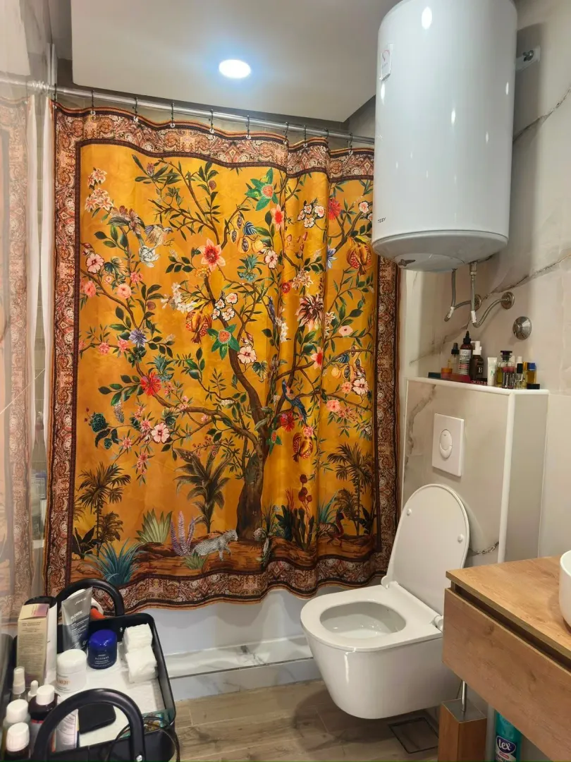 Manje kupatilo sa WC šoljom, bojlerom i tuš zavjesom sa cvjetnim dezenom.