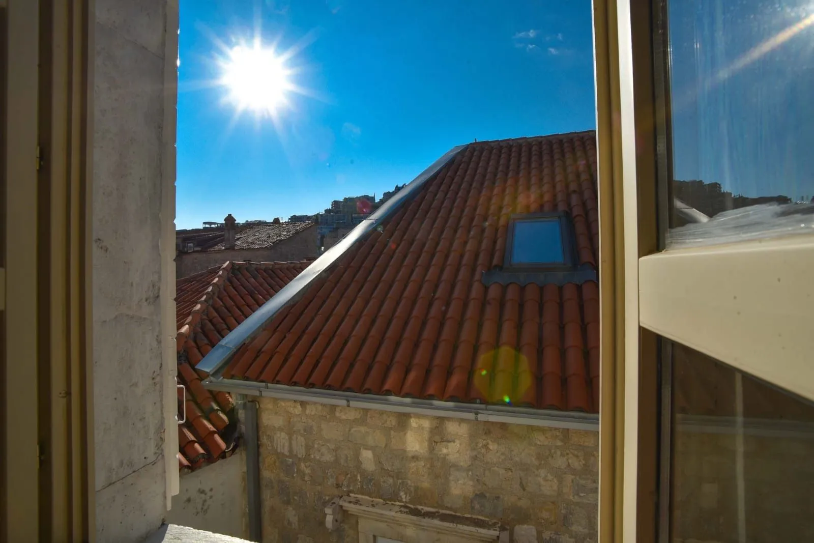 Pogled kroz prozor na krovove starih zgrada i sunčano nebo.