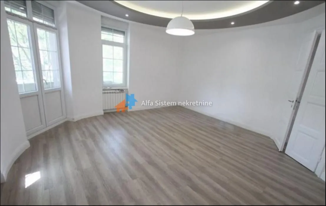 Prazna soba sa svetlim laminatom, belim zidovima i spuštenim plafonom sa rasvetom.