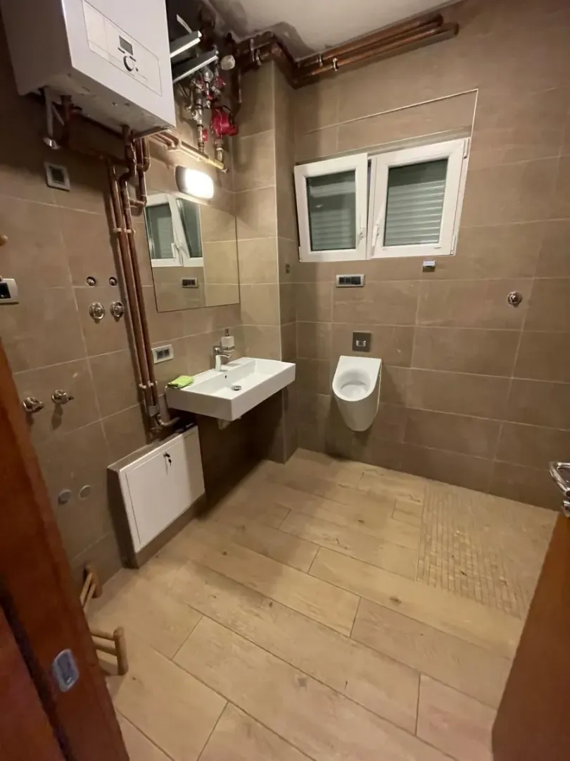 Kupatilo obloženo keramičkim pločicama sa umivaonikom, pisoarom i vidljivim instalacijama.