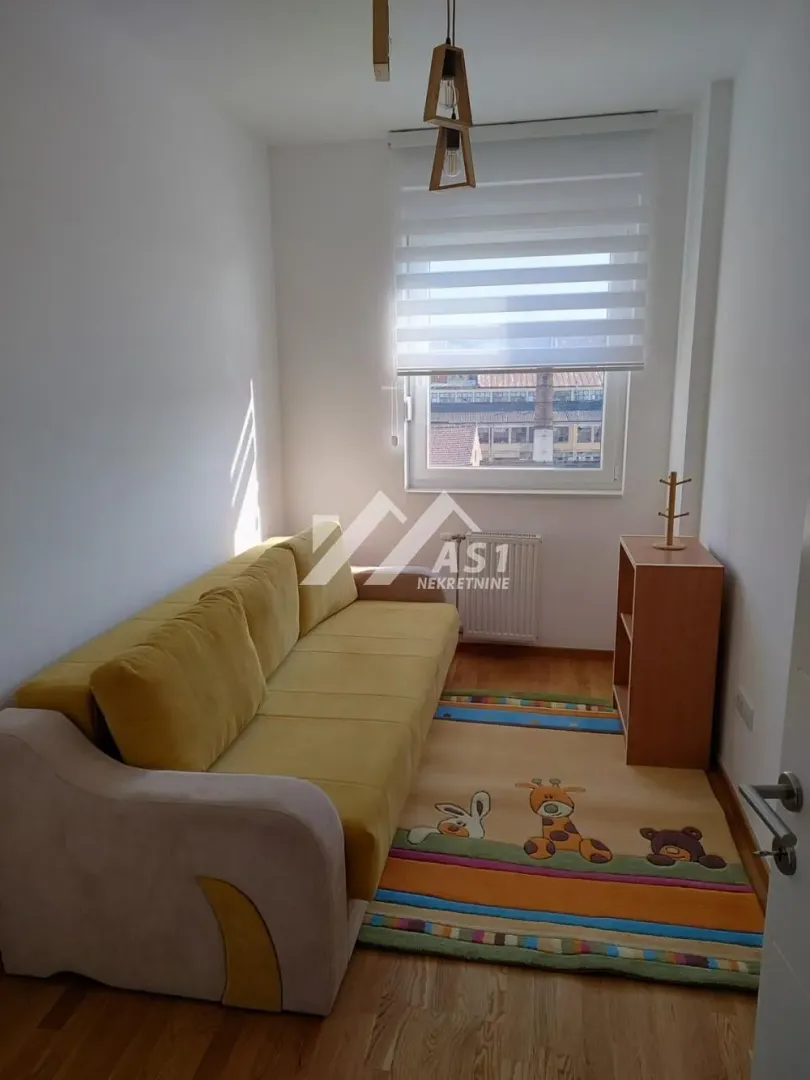 Manja dnevna soba sa žutim kaučem, tepihom sa dečijim motivima i prozorom.