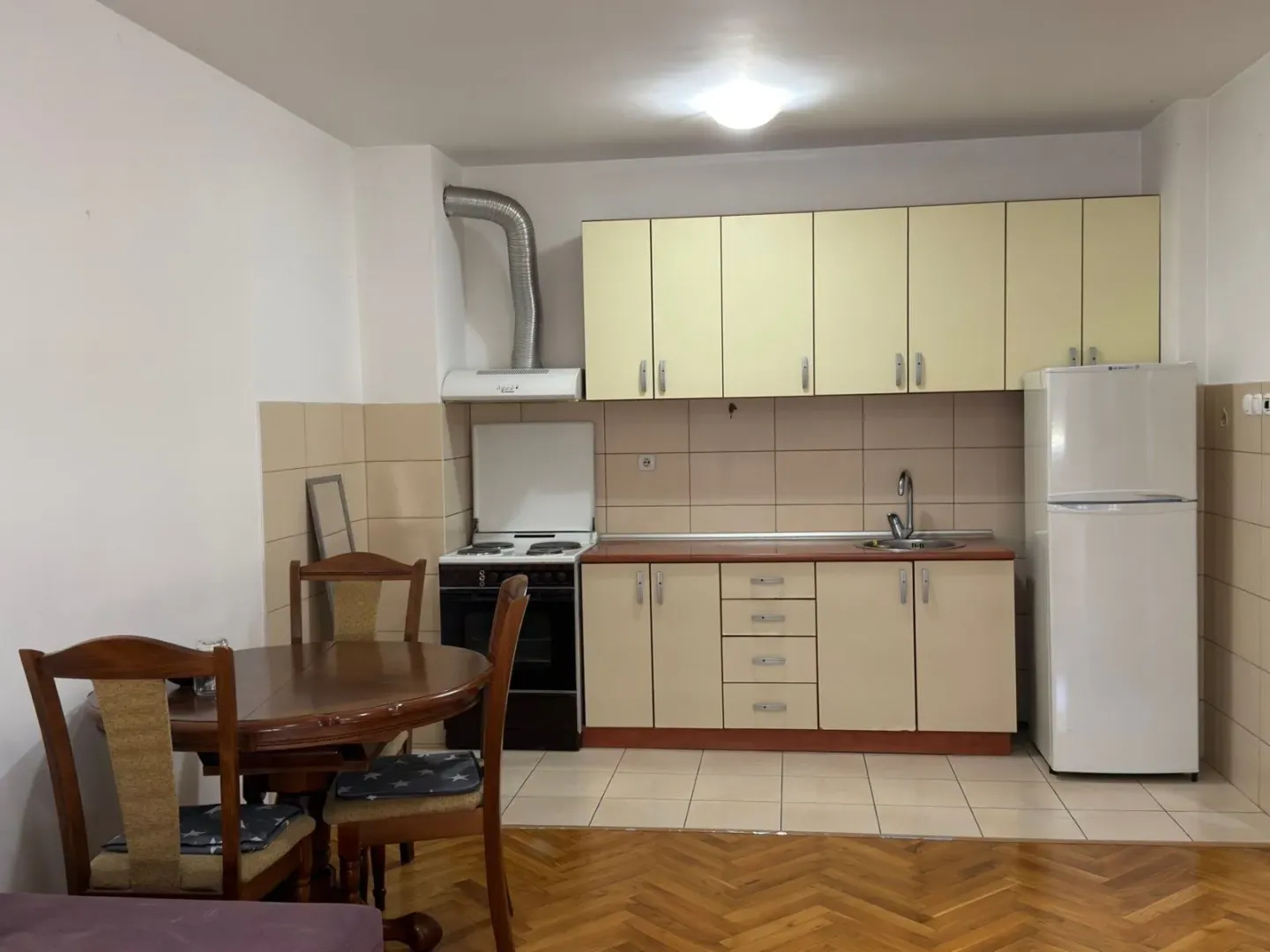 Kuhinja opremljena elementima, šporetom, frižiderom i trpezarijskim stolom sa stolicama.