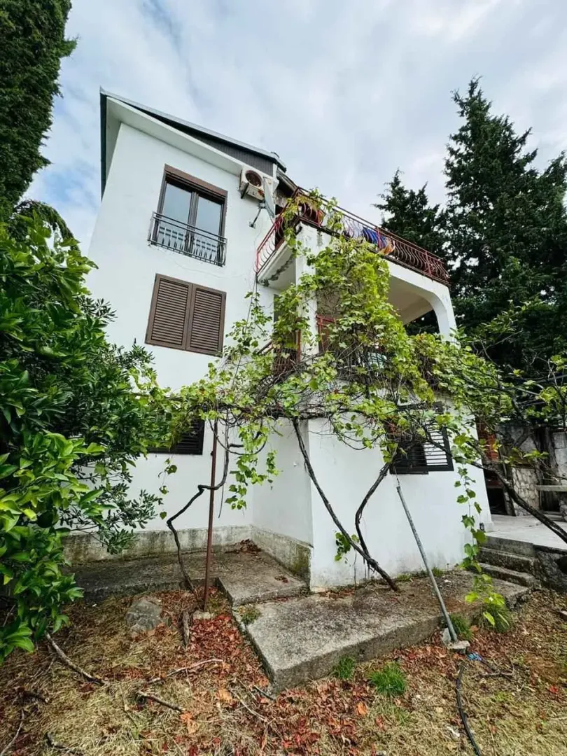 Pogled na spoljašnjost bijele kuće sa terasom i dvorištem sa zelenilom.