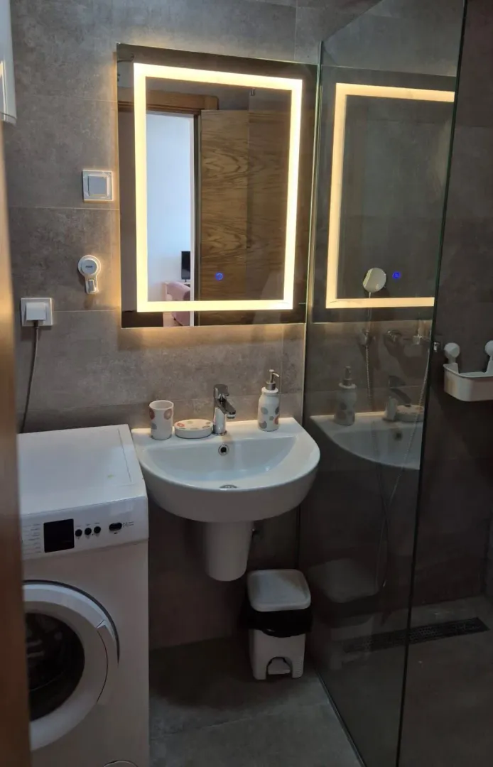 Bathroom with a washbasin, illuminated mirror, washing machine, and glass shower cabin.