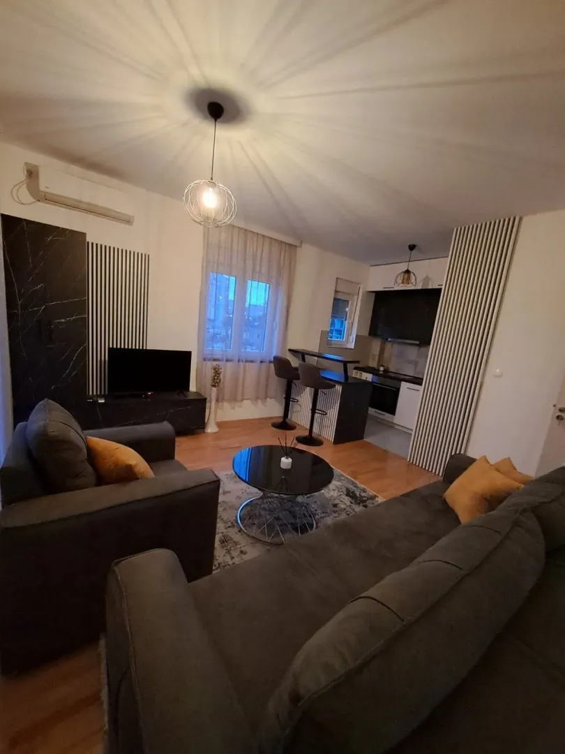 Bright open-plan living room with dark furniture and a kitchen area in the background.