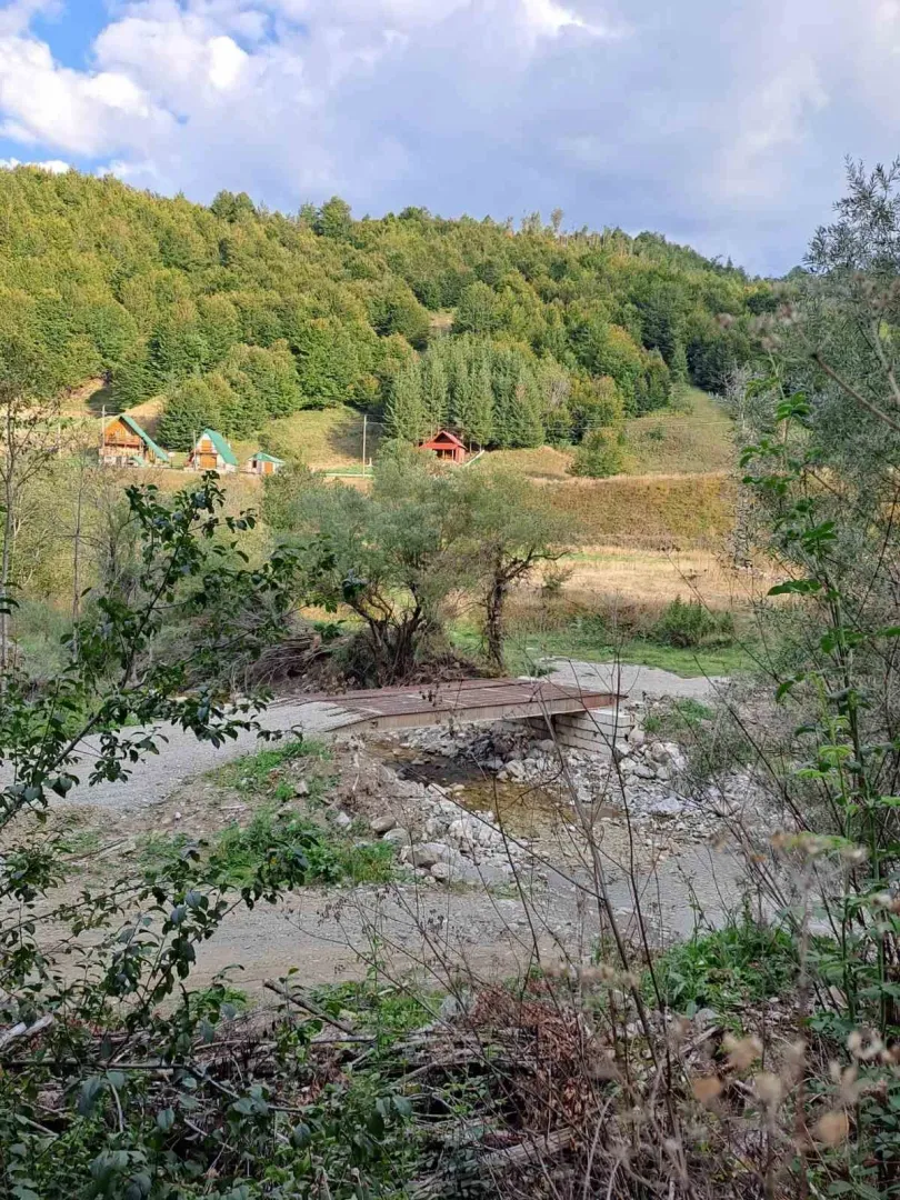 Pogled na brdoviti krajolik sa kućama, potokom i malim mostom.