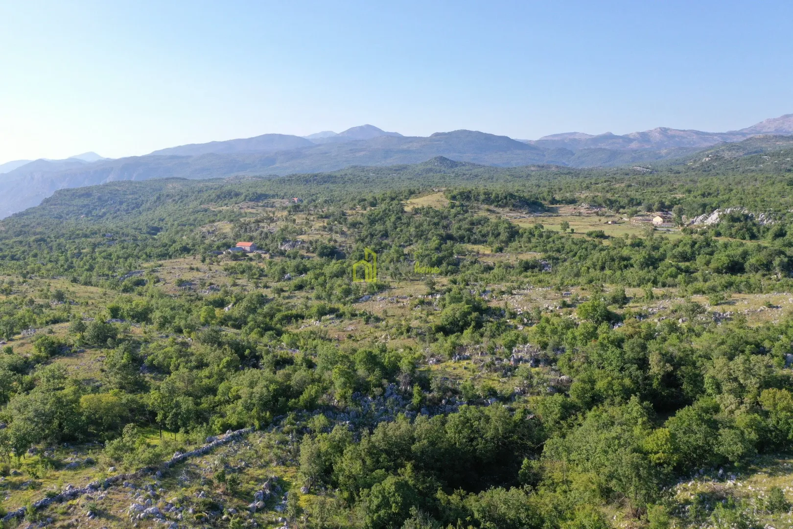 Pogled iz vazduha na brdoviti pejzaž sa šumom i planinama u pozadini.