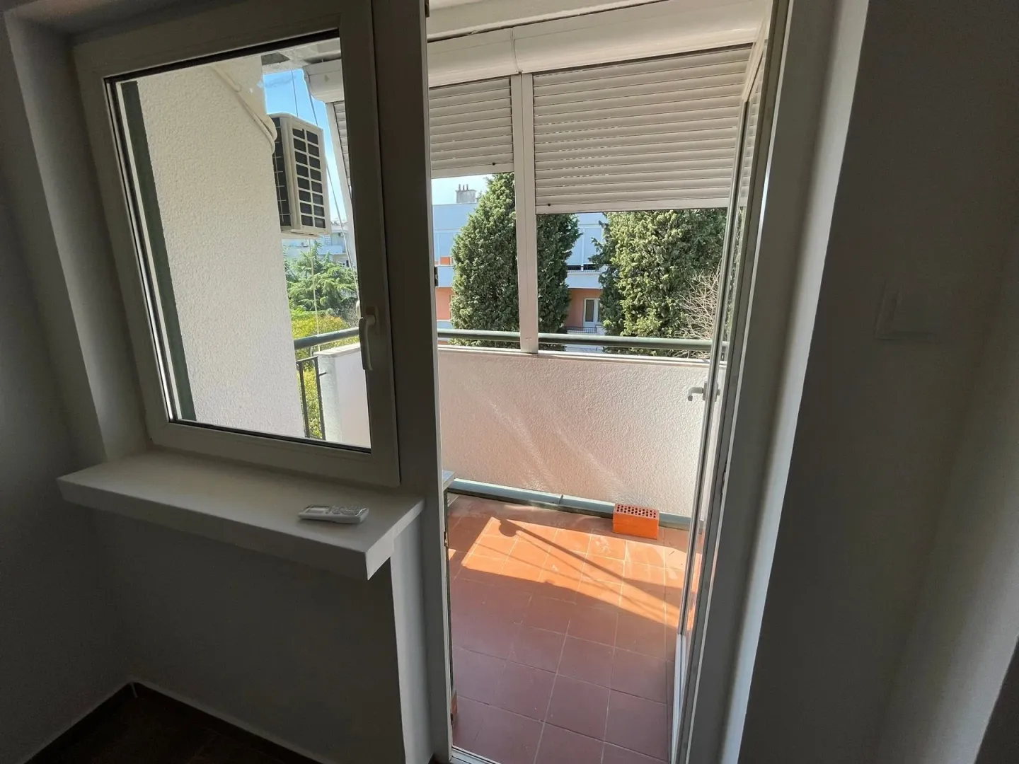 Access to a tiled balcony with a view of green areas and surrounding buildings.