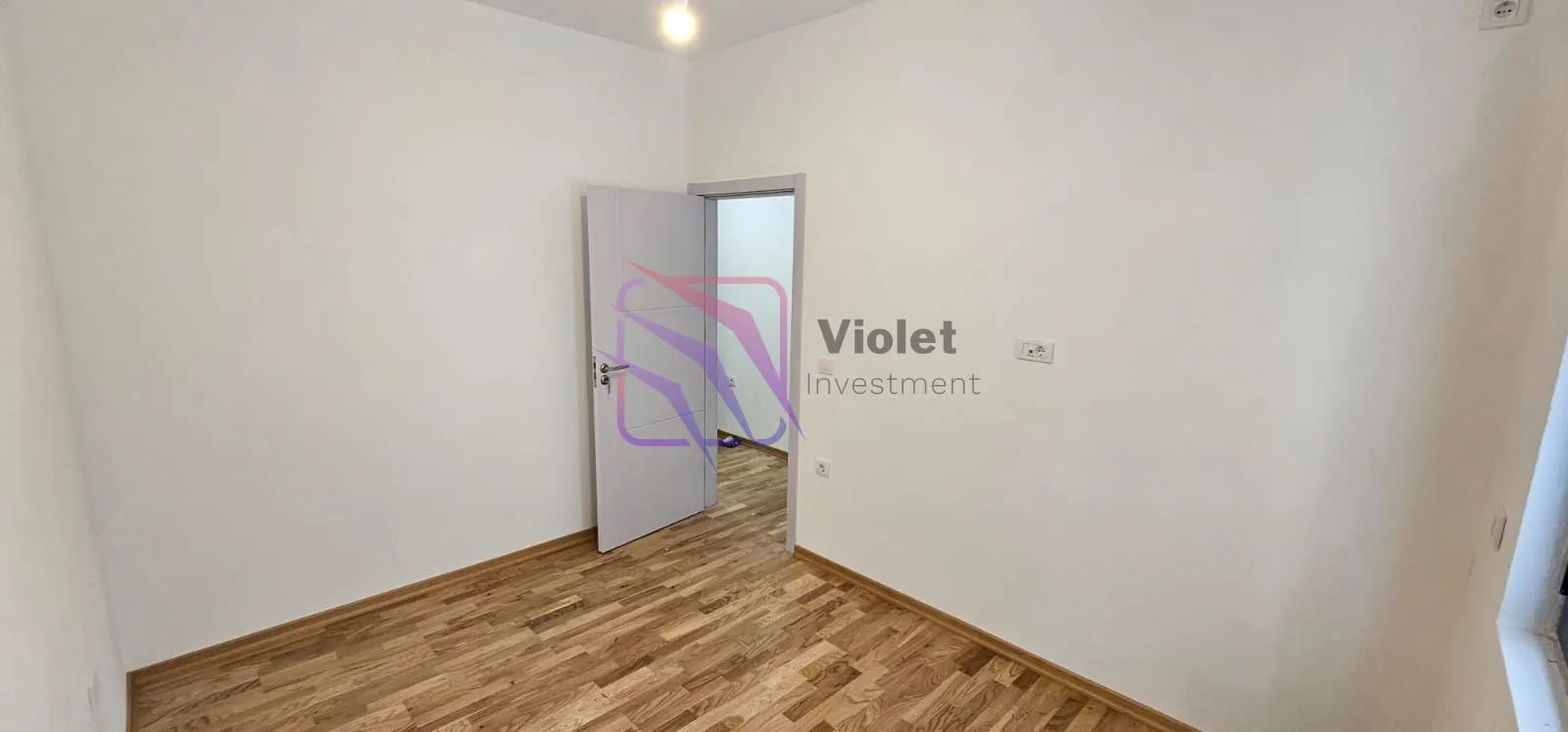 Prazna soba sa bijelim zidovima, drvenim podom i otvorenim vratima.