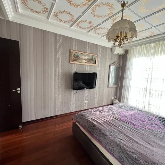 Furnished bedroom with a double bed, wall-mounted TV, and decorative ceiling.