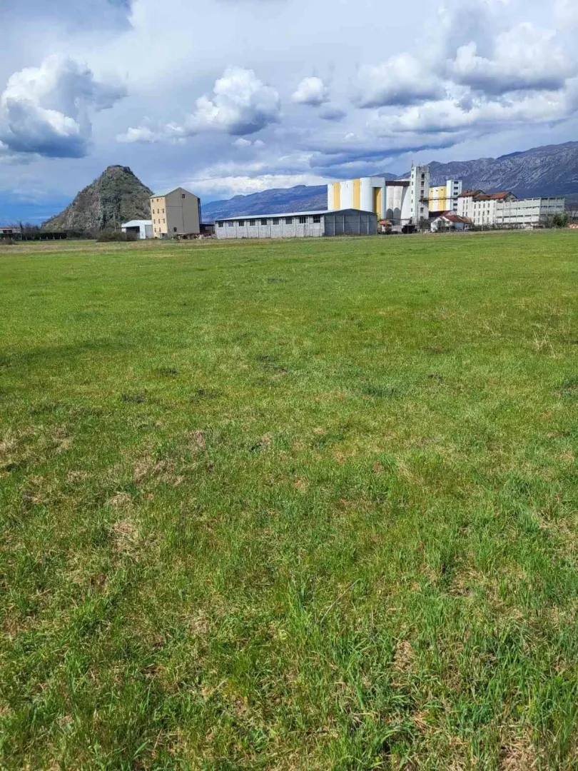 Prostrana zelena livada sa industrijskim objektima i planinama u pozadini.