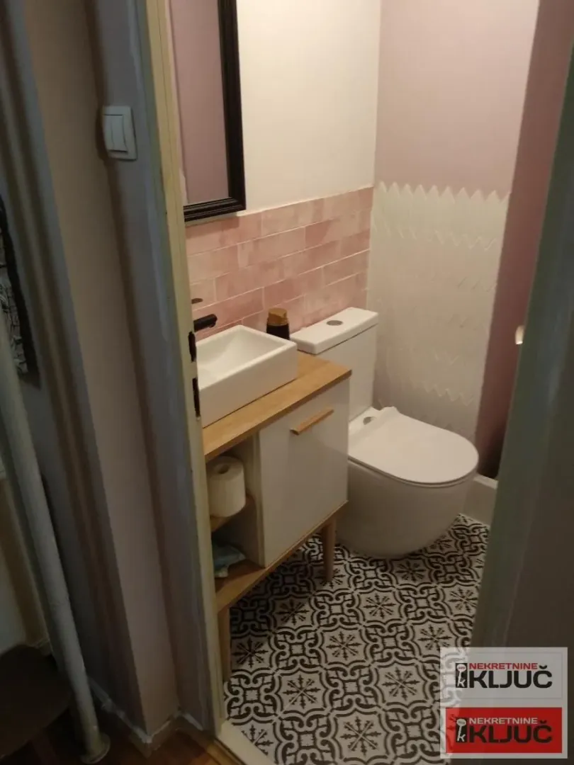 Manje kupatilo sa umivaonikom, toaletnom šoljom i dezeniranim pločicama na podu.