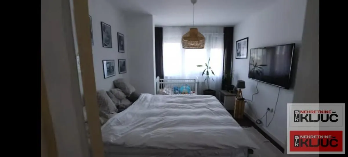 Prostrana spavaća soba sa velikim bračnim krevetom, TV-om na zidu i dečijim krevetcem u pozadini.