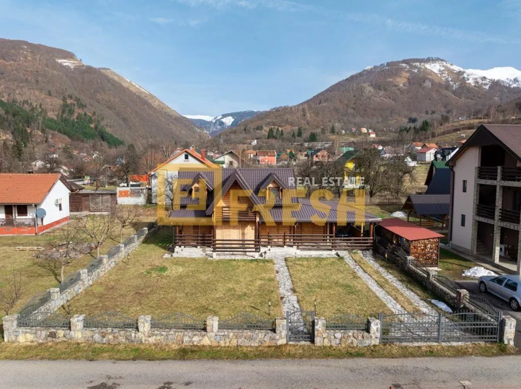 Pogled iz vazduha na drvenu kuću sa dvorištem i okolnim planinama.