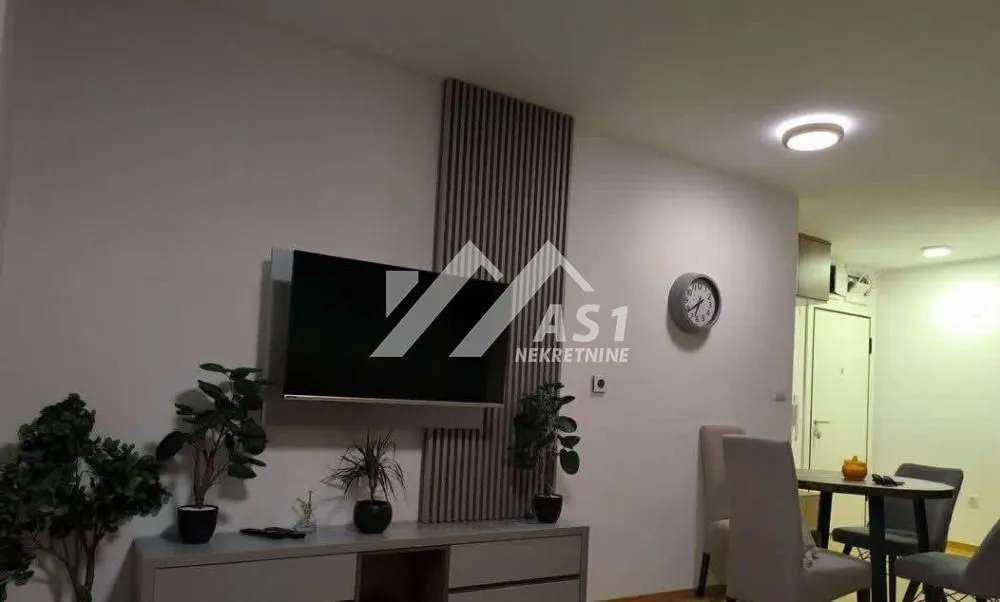 Dnevni boravak sa televizorom na zidu, komodom i trpezarijskim delom u pozadini.