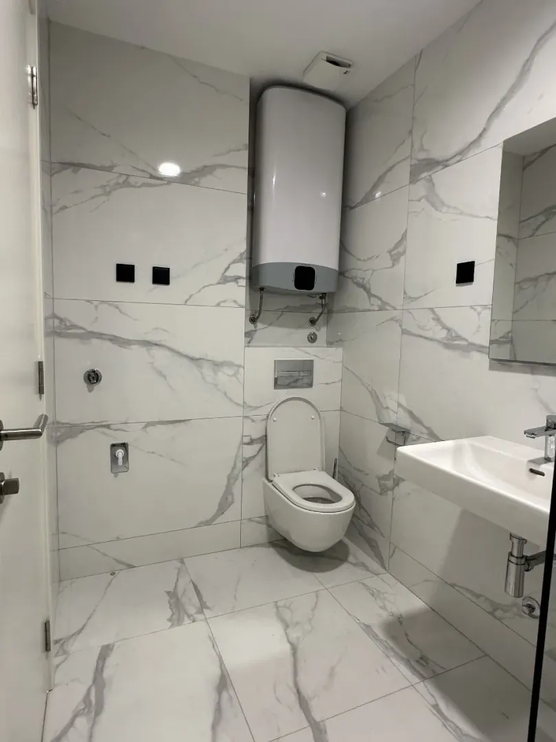 Moderno opremljeno kupatilo sa bijelim mermernim pločicama, wc šoljom, umivaonikom i bojlerom.