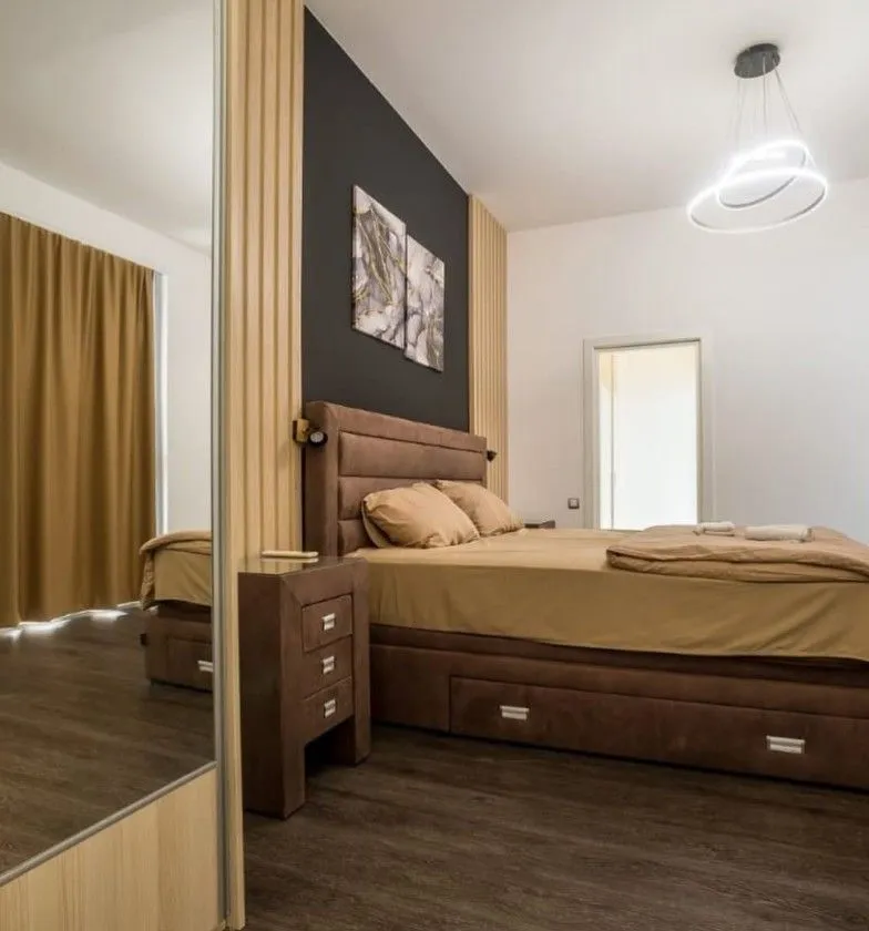 Moderna spavaća soba sa bračnim krevetom, noćnim stočićem i velikim ogledalom.