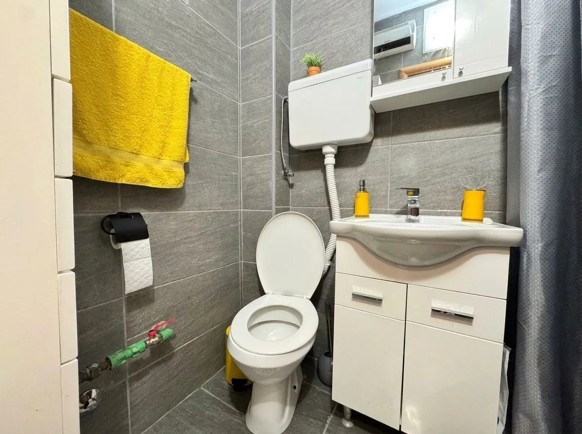 Manje kupatilo obloženo sivim pločicama sa toaletom, umivaonikom i žutim peškirom.