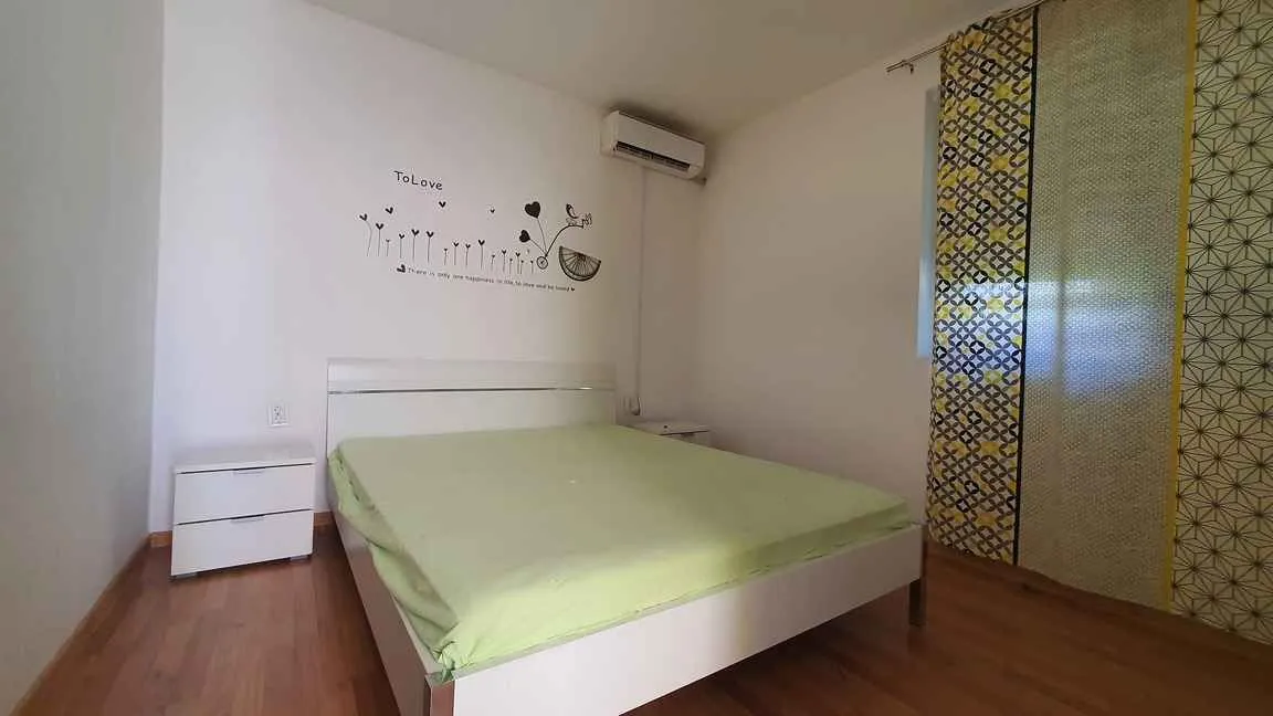 Spavaća soba sa bračnim krevetom, bijelim noćnim ormarićima i klimom na zidu.