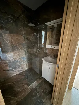 Kupatilo sa tuš kabinom obloženom tamnim mermernim pločicama i bijelim umivaonikom. - image 5