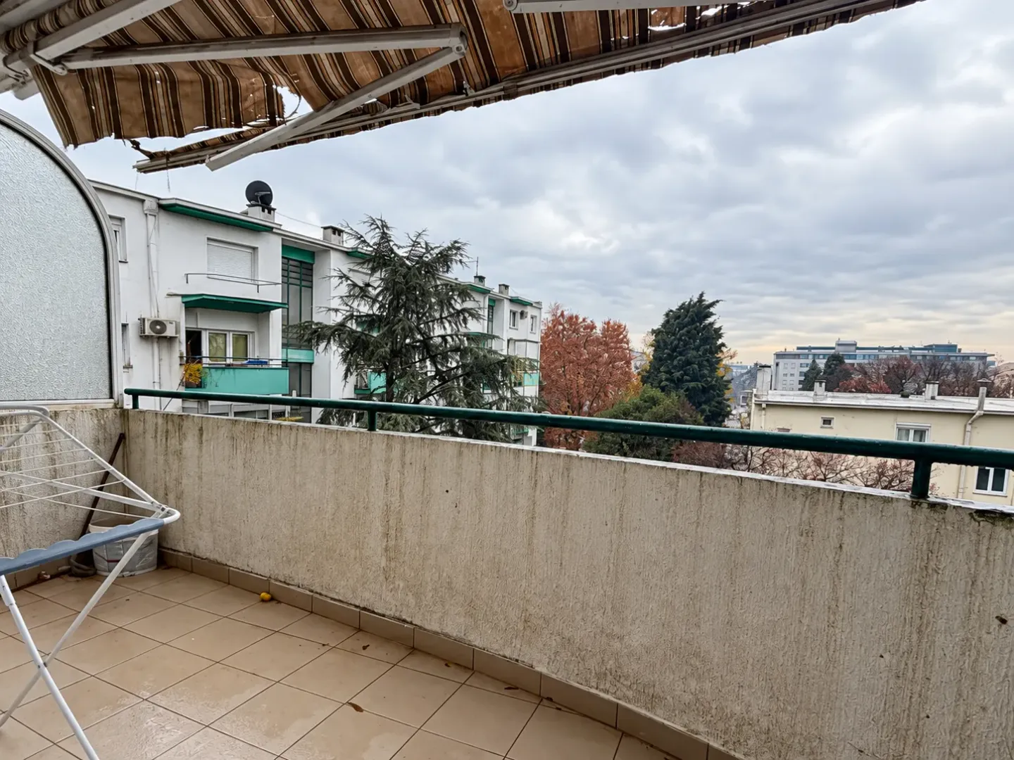 Natkriveni balkon sa pogledom na okolne zgrade i drveće.