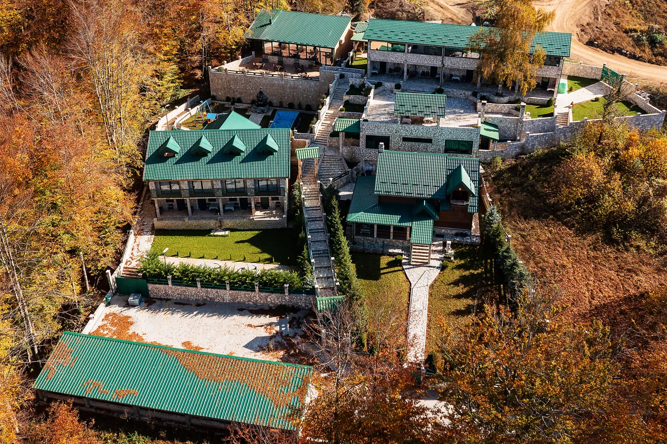 Pogled iz vazduha na kompleks kuća sa zelenim krovovima u šumskom okruženju.