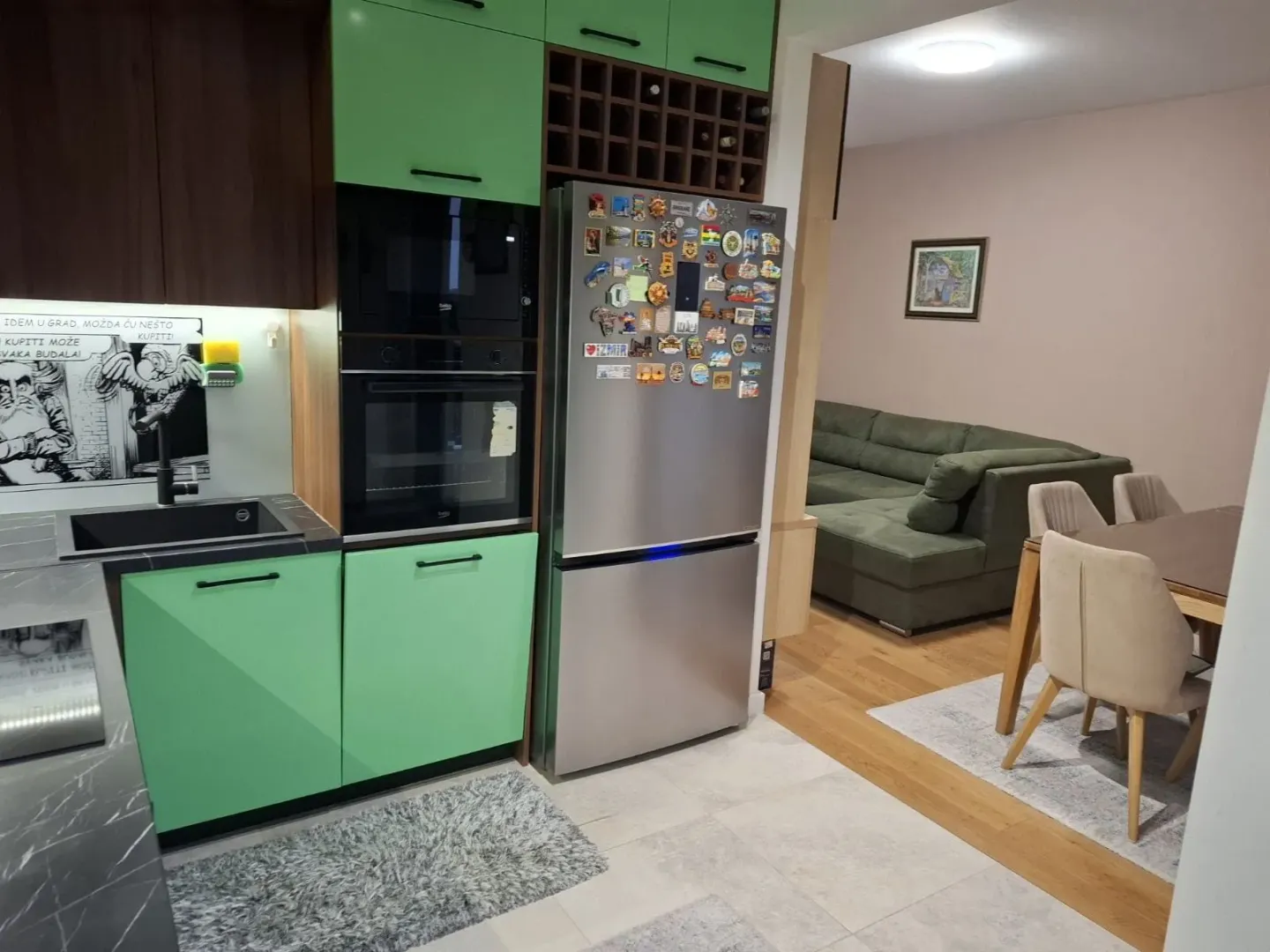 Moderna kuhinja u zelenim tonovima sa ugradnim aparatima i pogledom na trpezariju i dnevnu sobu.