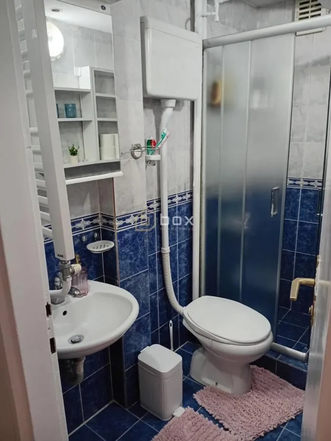 Manje kupatilo sa umivaonikom, WC šoljom i tuš kabinom obloženo pločicama.