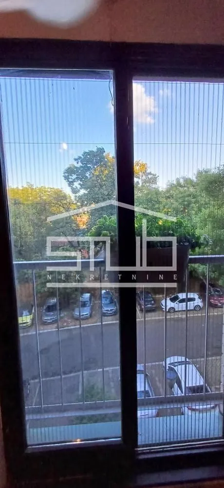 Pogled kroz prozor na okolno zelenilo i parking.