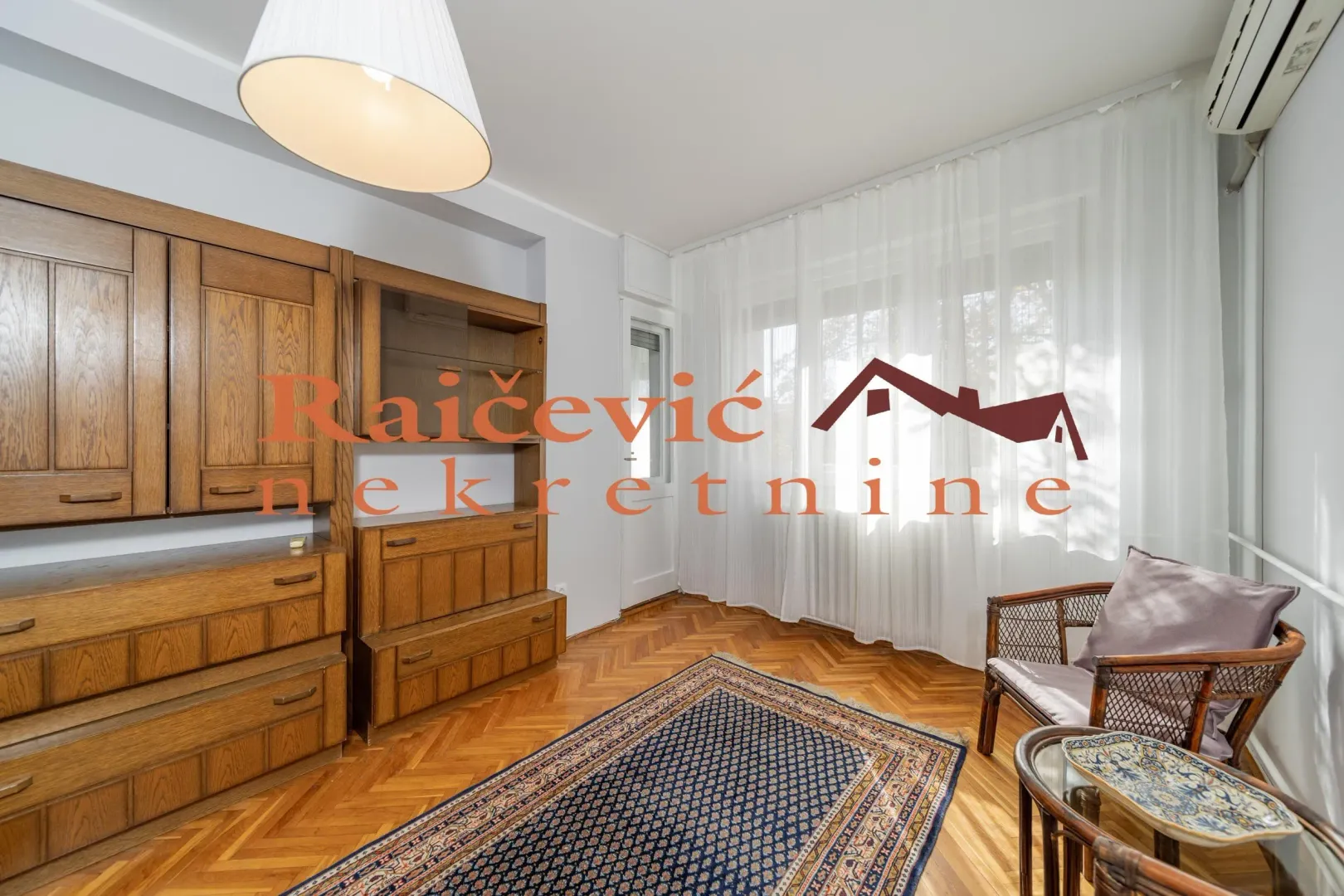 Living room with wooden furniture, parquet flooring, and a large window with white curtains.