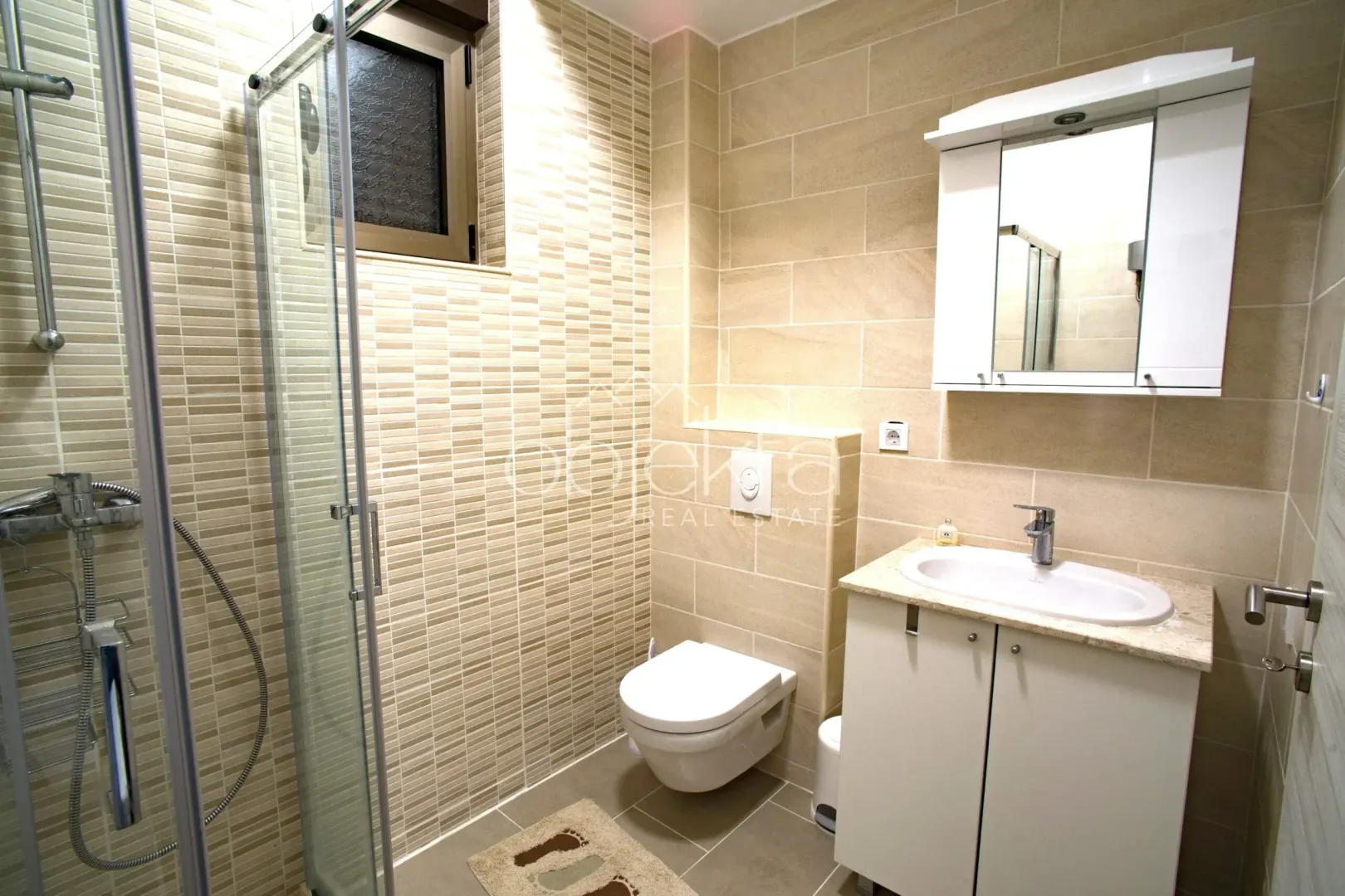 Kupatilo sa tuš kabinom, umivaonikom i ugradnom wc šoljom obloženo keramičkim pločicama.