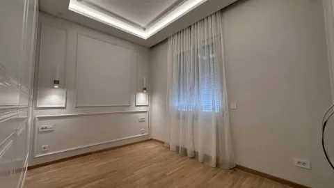 Prazna soba sa drvenim podom, dekorativnim lajsnama na zidu i prozorom sa belim zavesama. - image 5