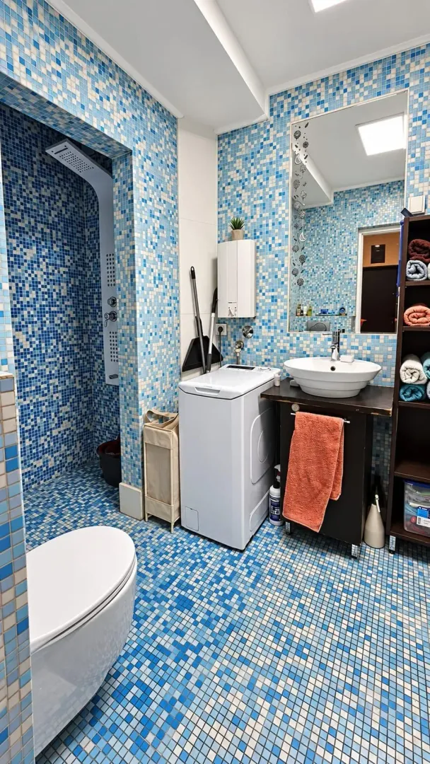 Bathroom lined with blue mosaic tiles featuring a shower cabin, washbasin, and washing machine.