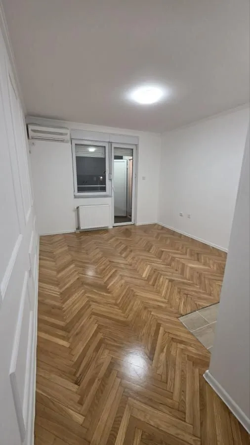 Prazna soba sa drvenim parketom, belim zidovima i klima uređajem.