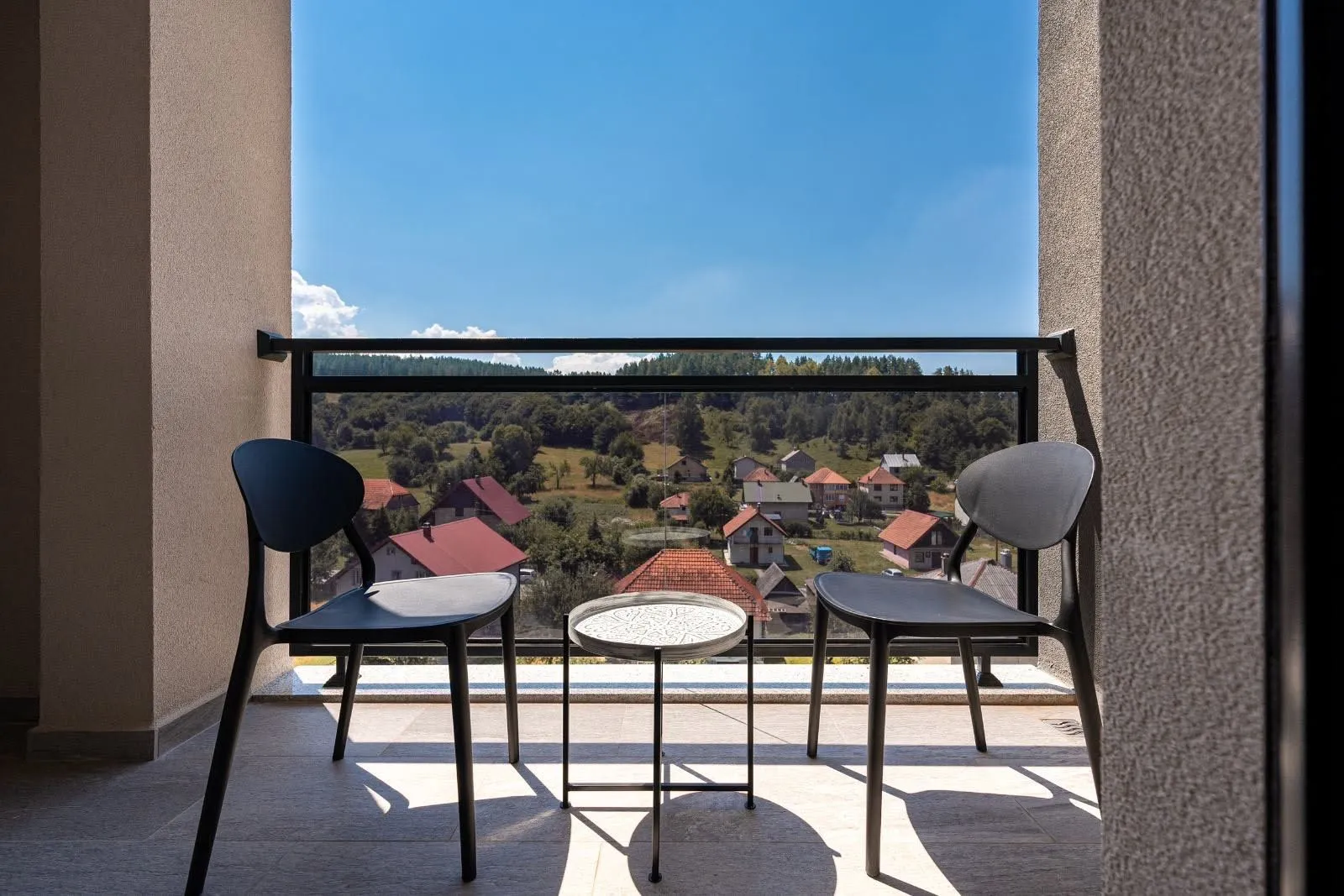 Natkriveni balkon sa dvije crne stolice i stočićem i pogledom na prirodu i naselje.