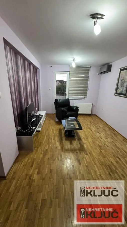 Dnevna soba sa drvenim podom, foteljom i TV komodom.