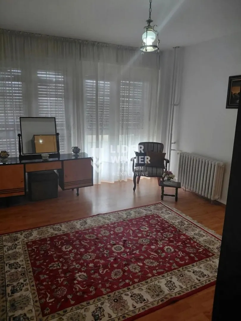 Spavaća soba sa velikim prozorom, tepihom i komodom sa ogledalom.