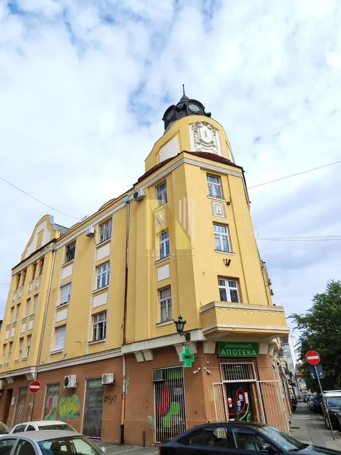 Eksterijer višespratne žute stambeno-poslovne zgrade sa apotekom u prizemlju.
