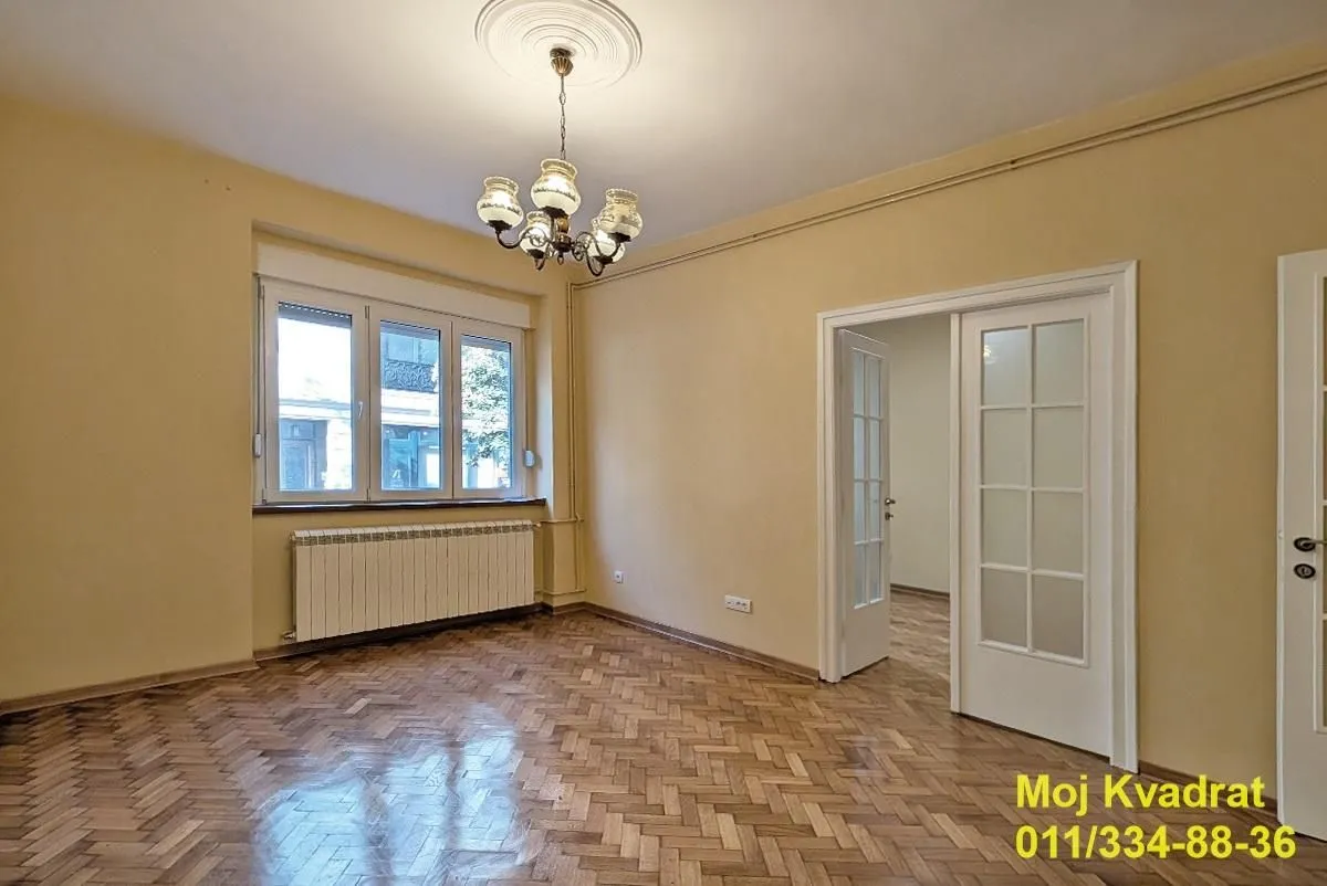 Prazna dnevna soba sa parketom, lusterom i vratima koja vode u drugu prostoriju.
