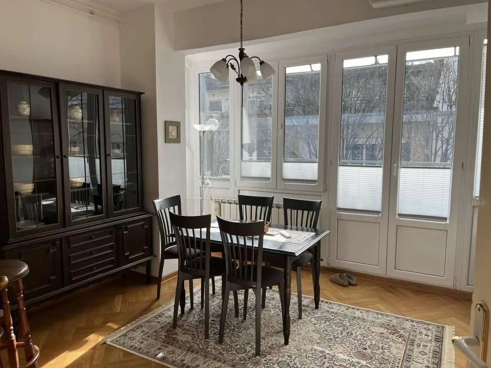 Dining area with a dark wooden table, chairs, and a cabinet next to large windows.