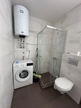Smaller bathroom equipped with a shower cabin, toilet, washing machine, and water heater. - image 6