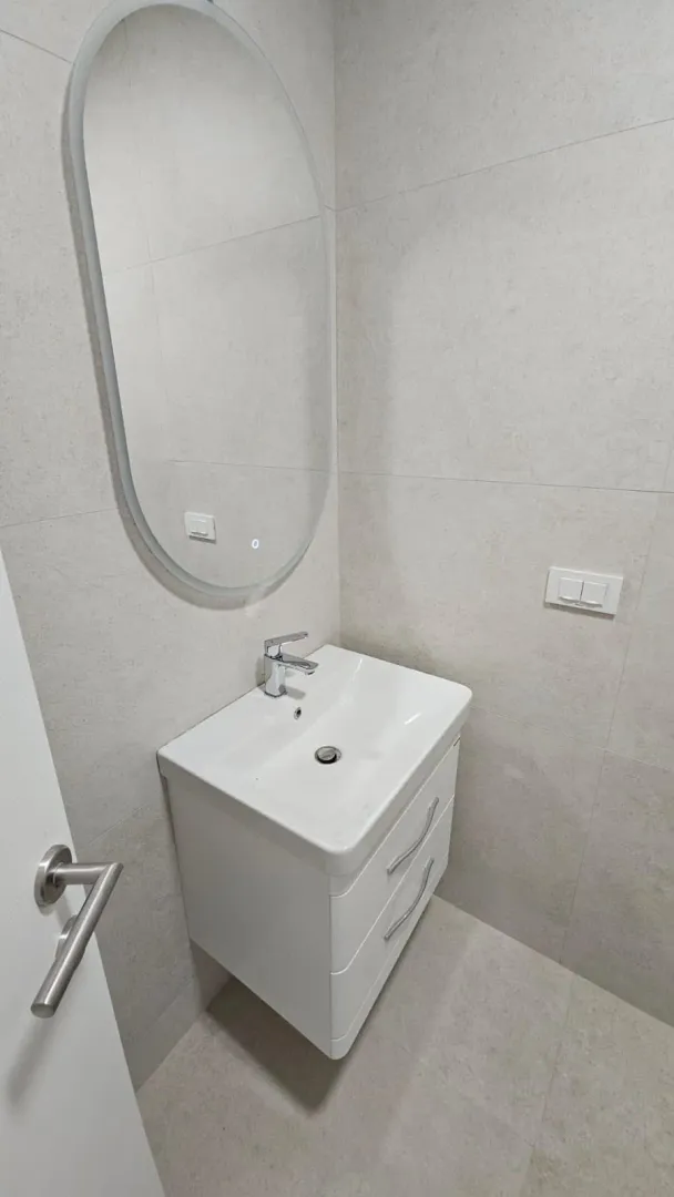 Savremeno opremljeno kupatilo sa umivaonikom, ogledalom i pločicama u neutralnim tonovima.